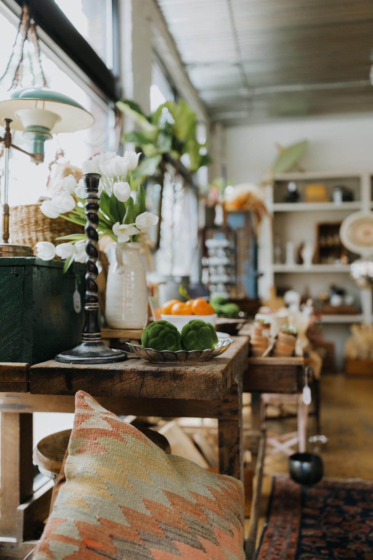 Interior of a cozy room with a wooden table displaying fresh vegetables, including green broccoli and orange tomatoes, next to flower vases, with shelves in the background.
