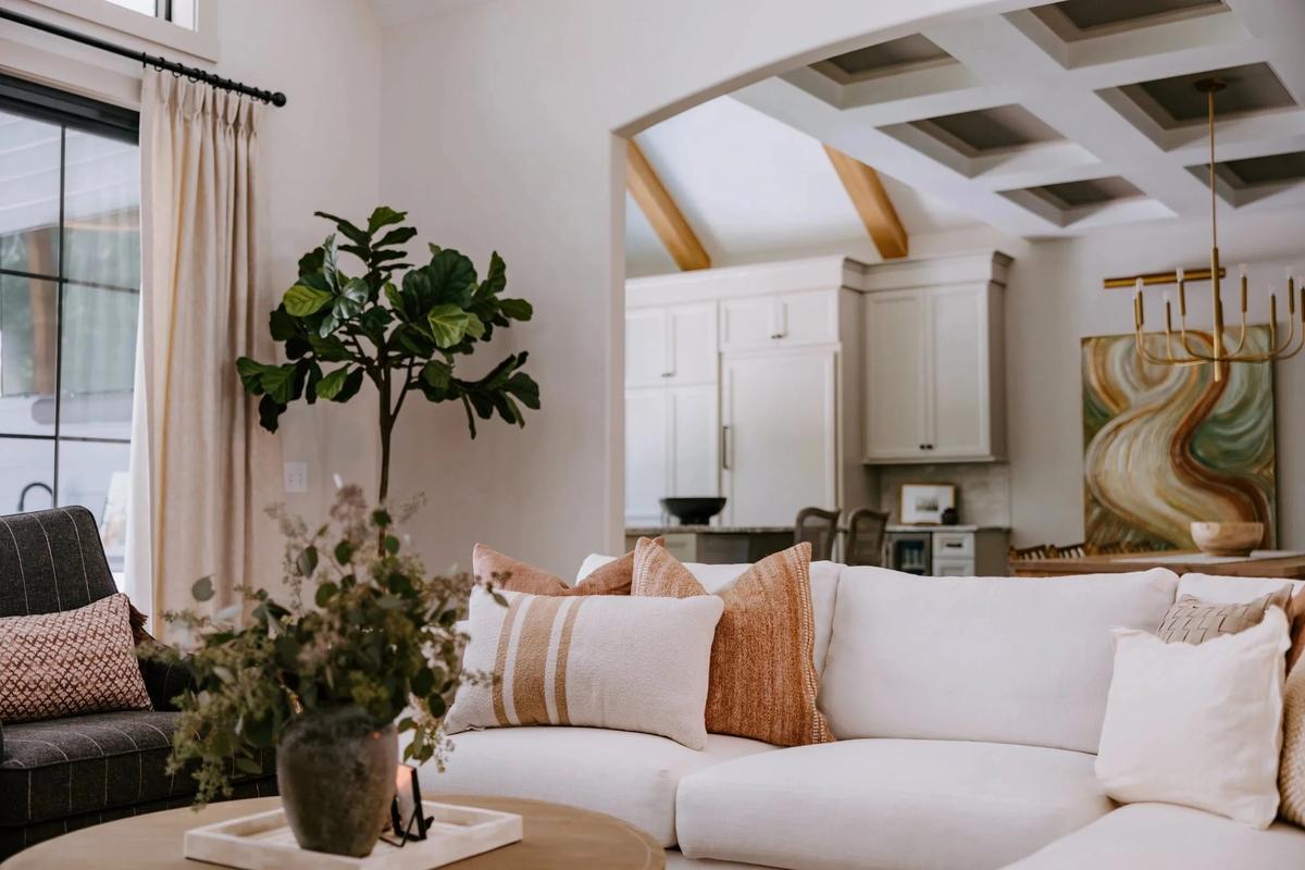 A cozy living room with a white sectional sofa decorated with beige and rust-colored pillows, a wooden coffee table with a flower vase, a potted plant near a window with cream curtains, and a view of a kitchen area with white cabinets and modern art on the wall.