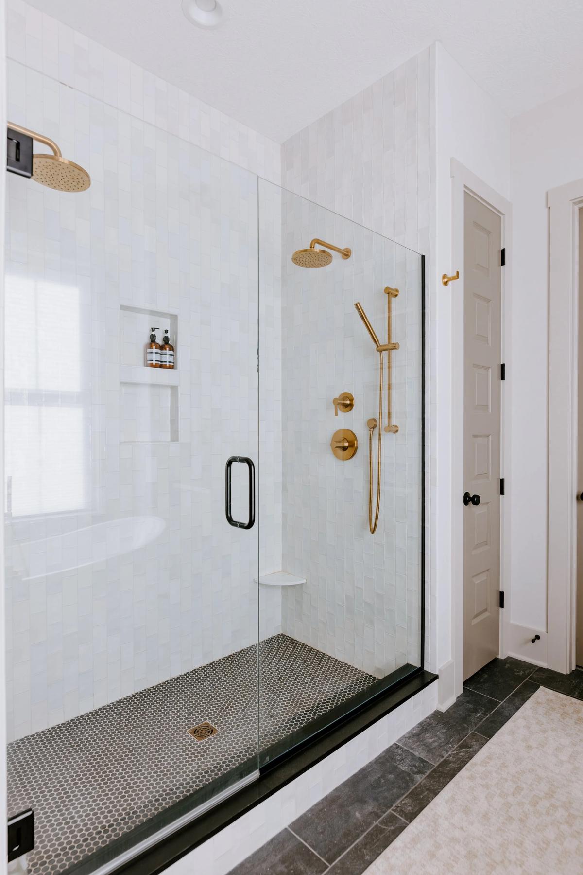 Modern glass-enclosed shower with gold fixtures, a built-in shelf with soap bottles, white tiled walls, and a dark tile floor.