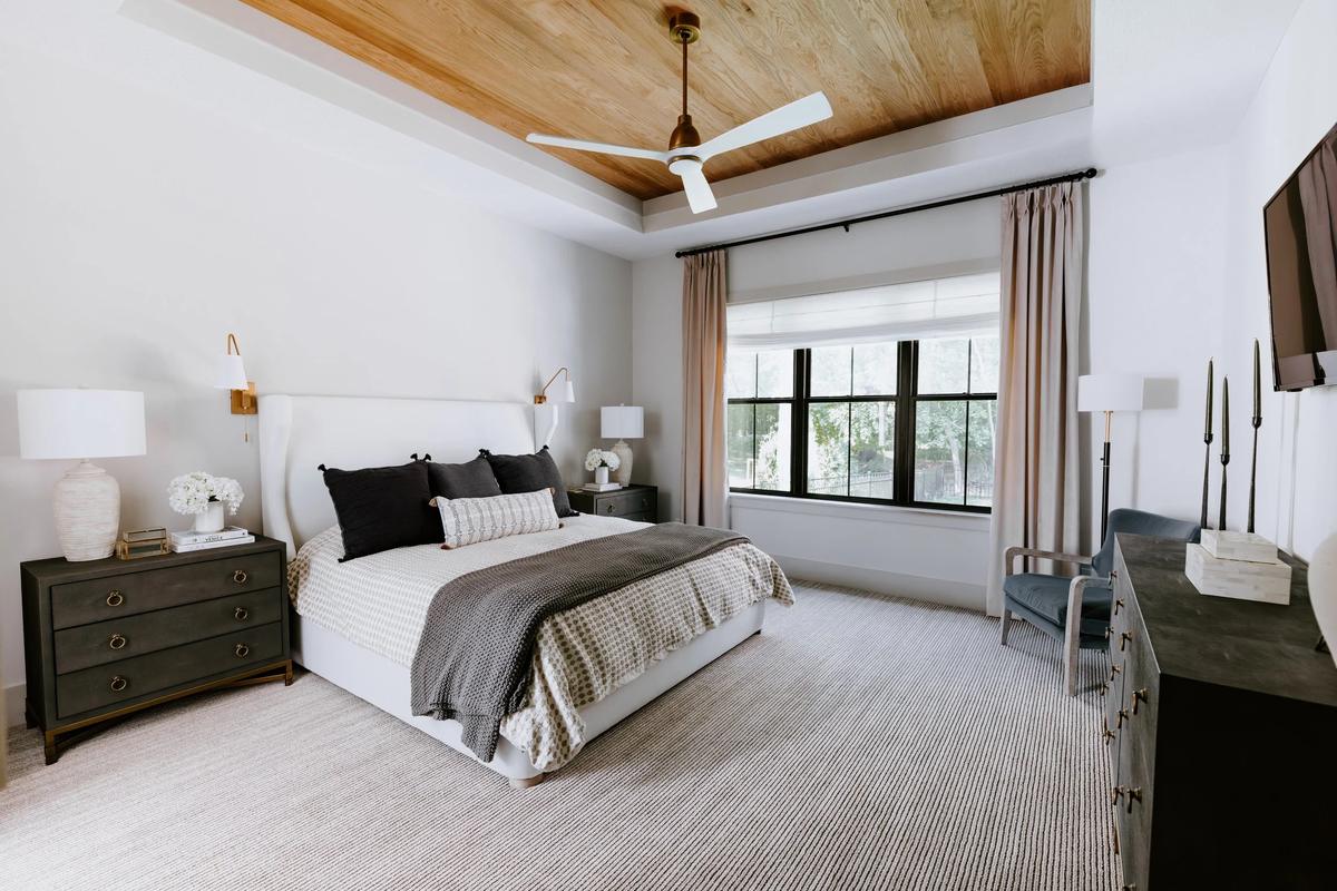 A modern bedroom with a large window, white walls, wood ceiling, gray nightstands, a bed with black and white pillows, lamps, a ceiling fan, a chair, and a dresser.