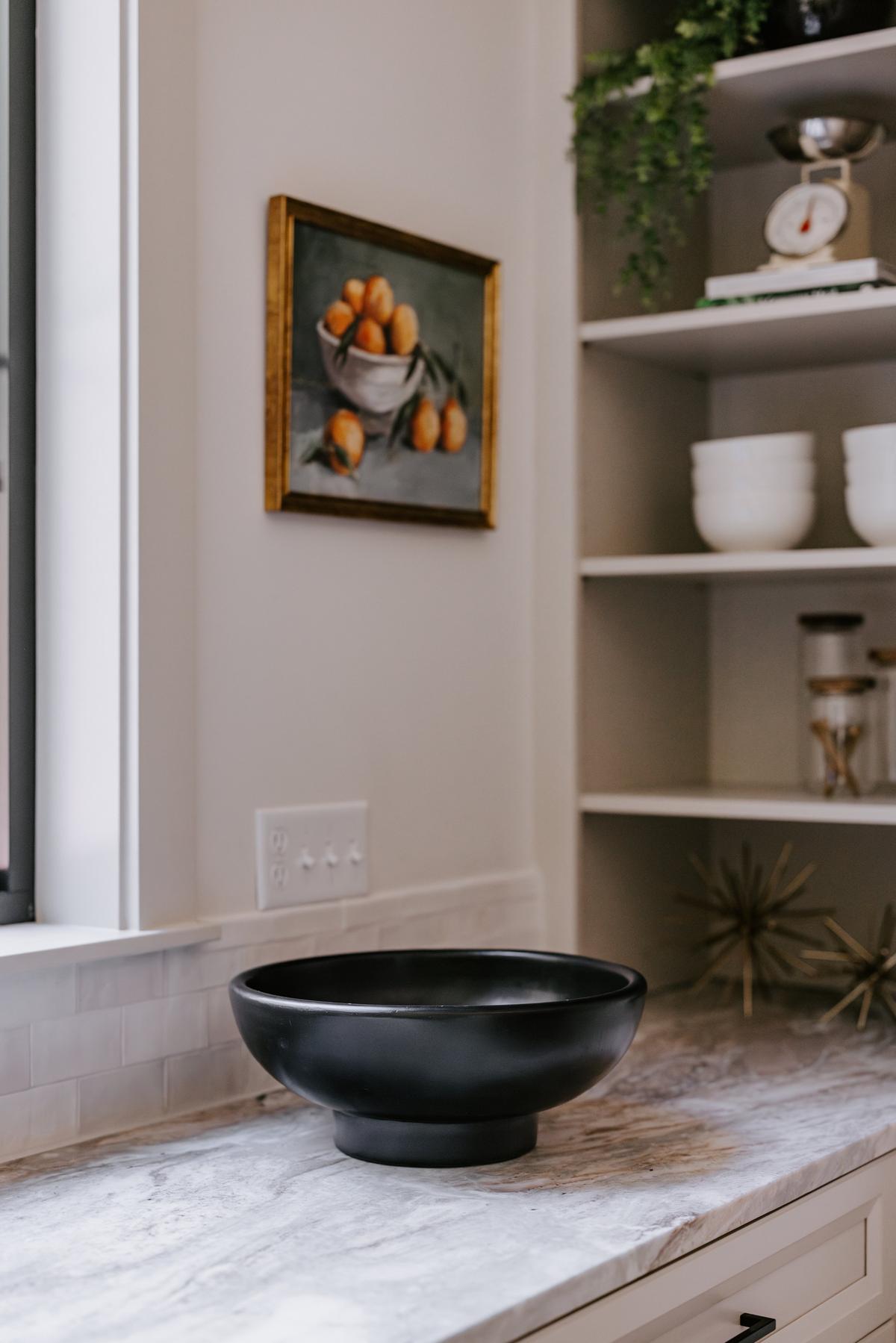 Black bowl on a marble kitchen countertop with a painting of oranges in a bowl on the wall behind and shelves with white bowls and a scale.