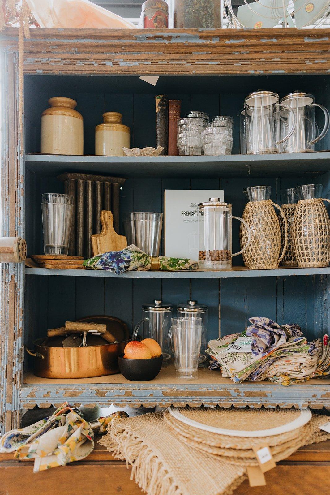 Open wooden cabinet filled with glassware, cups, bowls, books, and decorative items, with a stack of woven placemats and cloth napkins in front.