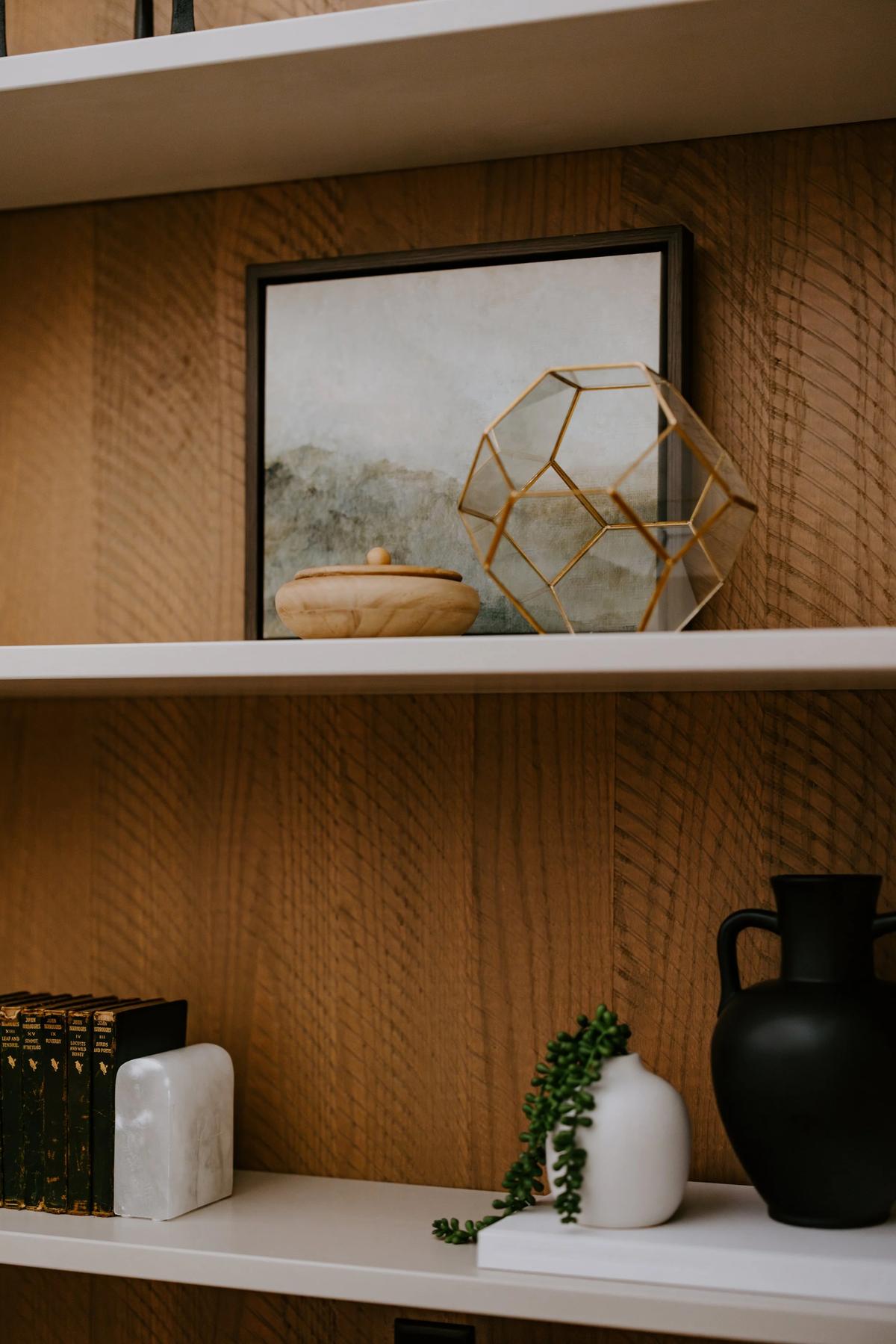 Decorative shelves with a framed art piece, a geometric gold sculpture, a wooden container, a white vase with a green trailing plant, a black vase, and a set of black books with gold lettering.