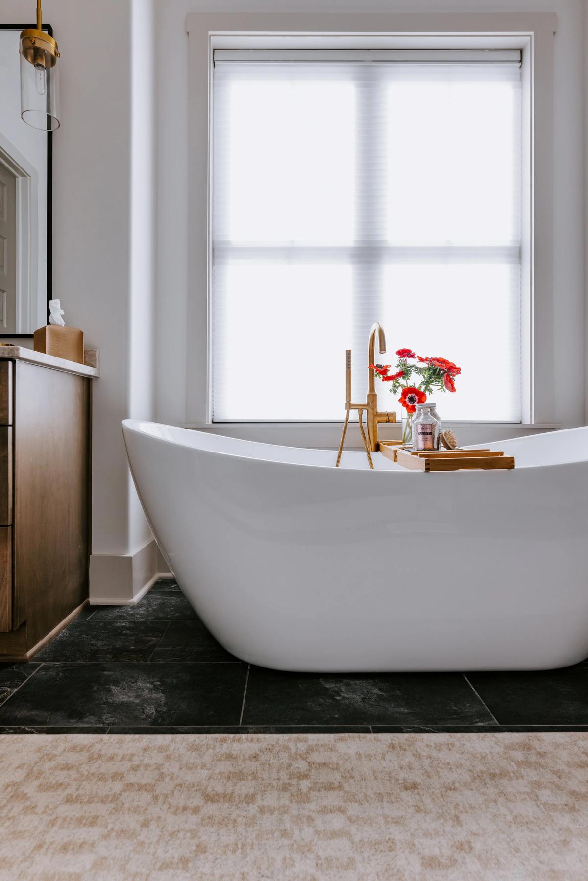 A modern bathroom with a white freestanding bathtub with a wooden tray and red flowers, positioned in front of a large window with white blinds, black floor tiles, and a beige area rug.