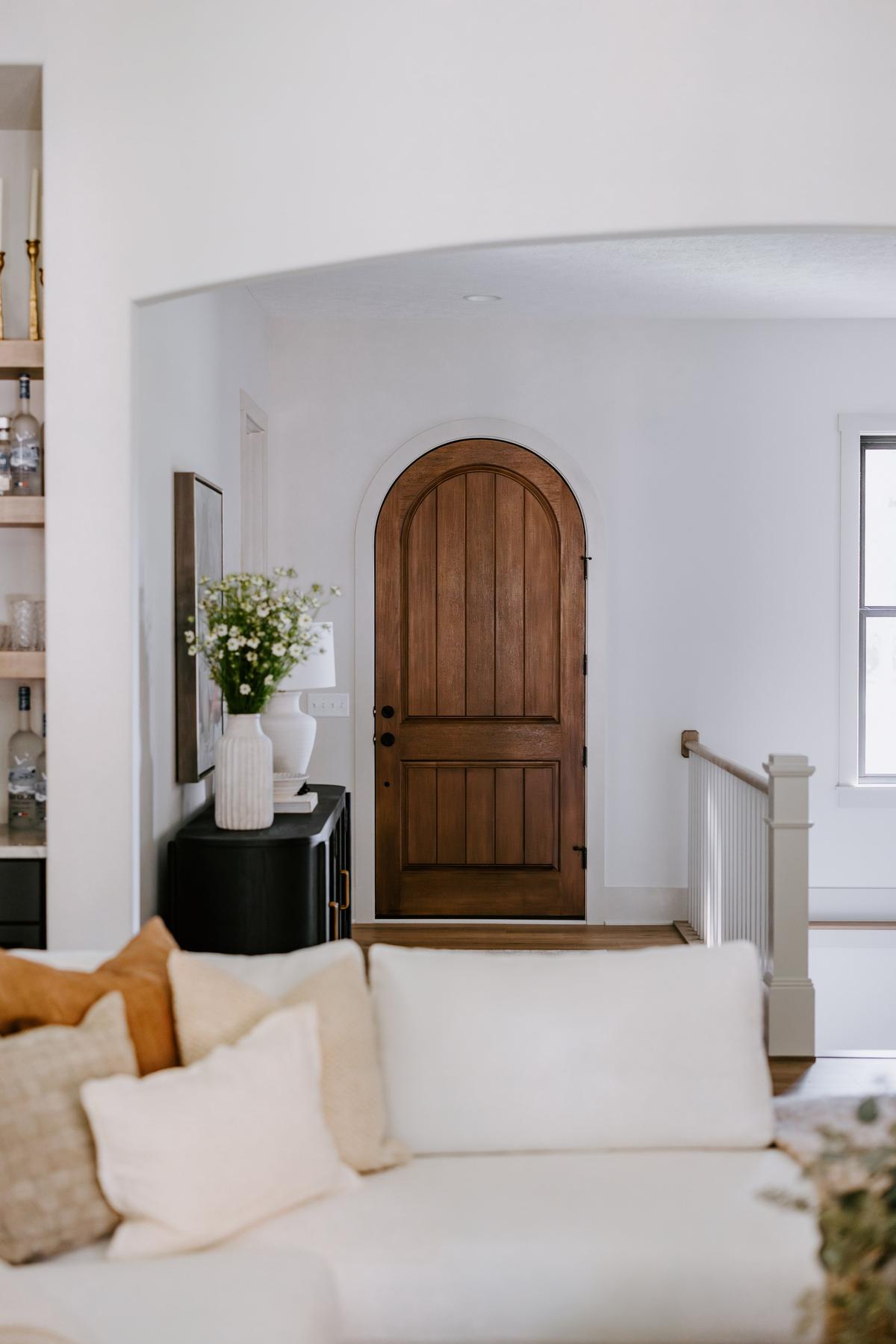 View of a cozy living room with a closed wooden arched door, white walls, a black console table with vases and flowers, a framed picture, and a sofa with beige and brown pillows in the foreground.
