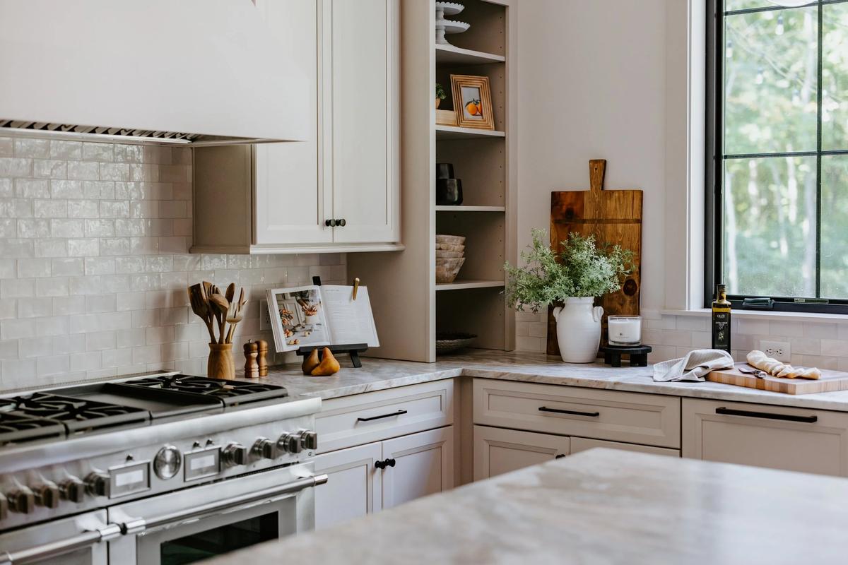Kitchen with white cabinets, marble countertops, a stove, open shelving with decorative items, a window with trees outside, and kitchen accessories like a vase with greenery, cutting board, and utensils.