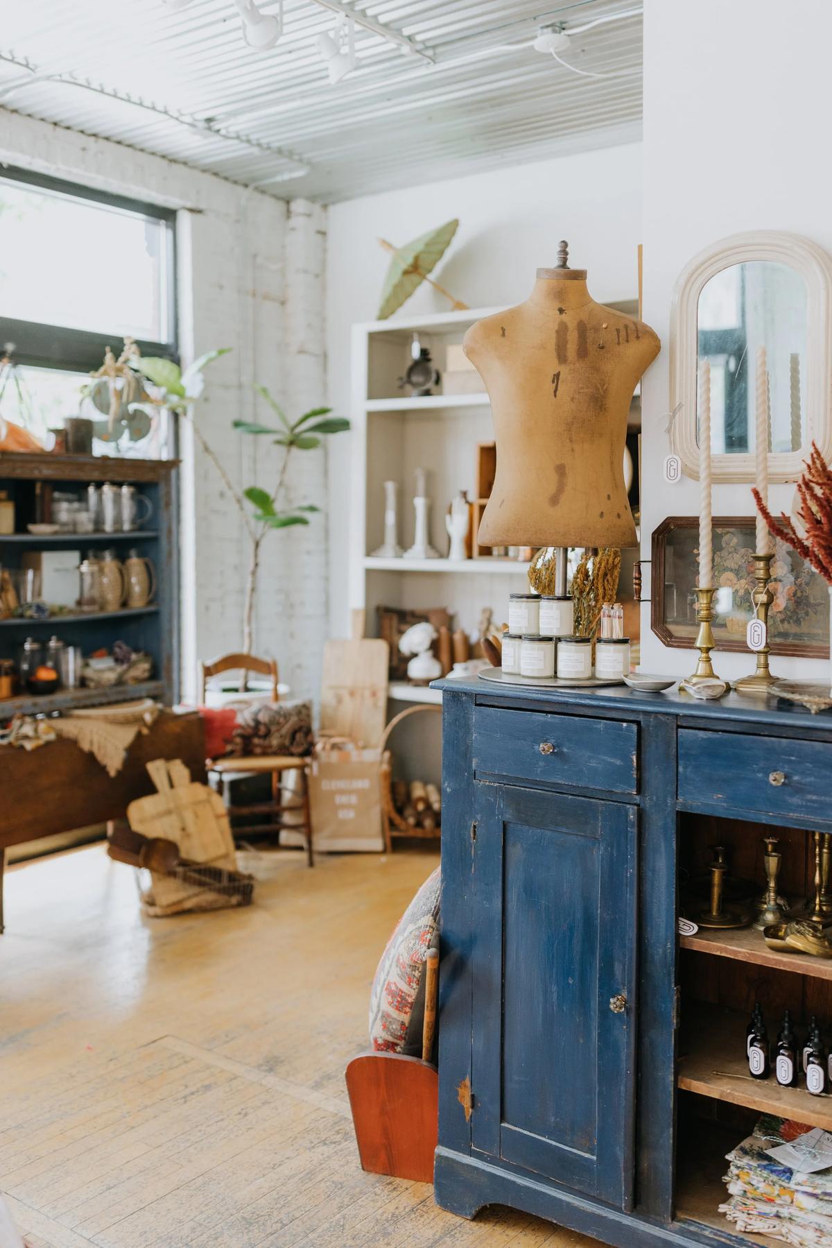 Interior of a vintage store with a mannequin torso on a blue distressed cabinet, shelves with various decorative items, and a window letting in natural light.