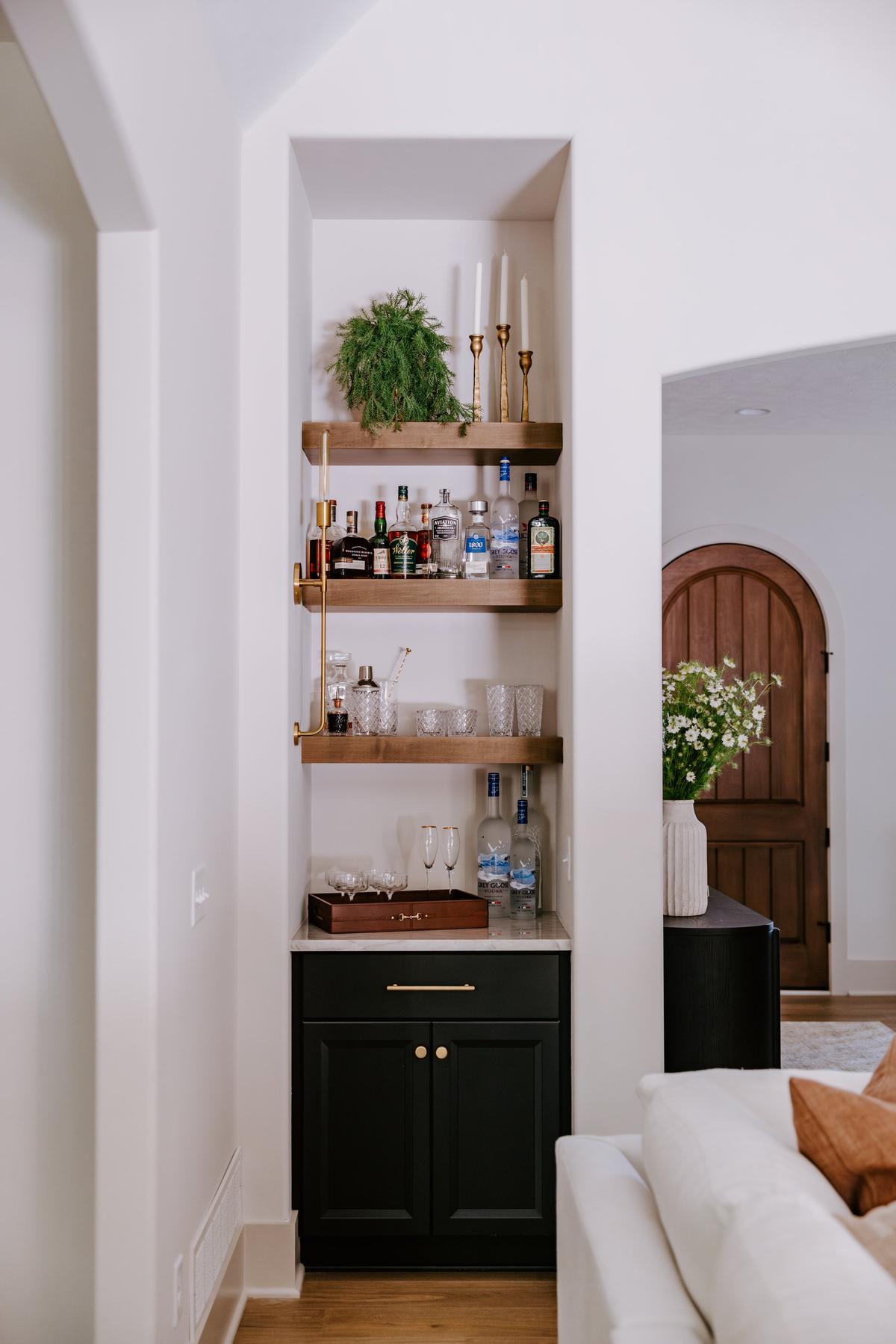 A built-in bar with three wooden shelves filled with liquor bottles, glassware, and candles, and a black cabinet with gold details beneath, located in a corner of a living room with a white wall and a doorway with a wooden door in the background.