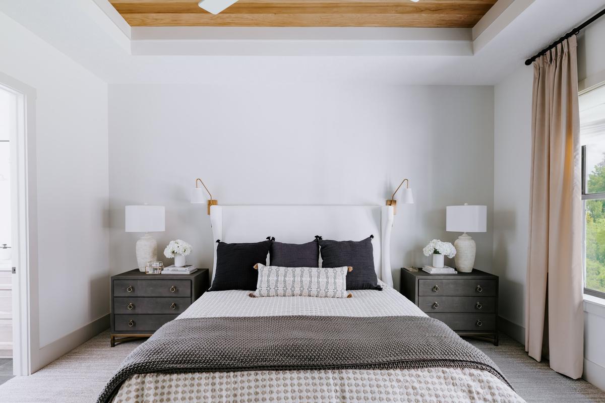 A neatly made bedroom with a white upholstered headboard, six black and patterned pillows, two gray nightstands with white lamps, and a large window with beige curtains in a modern white room.
