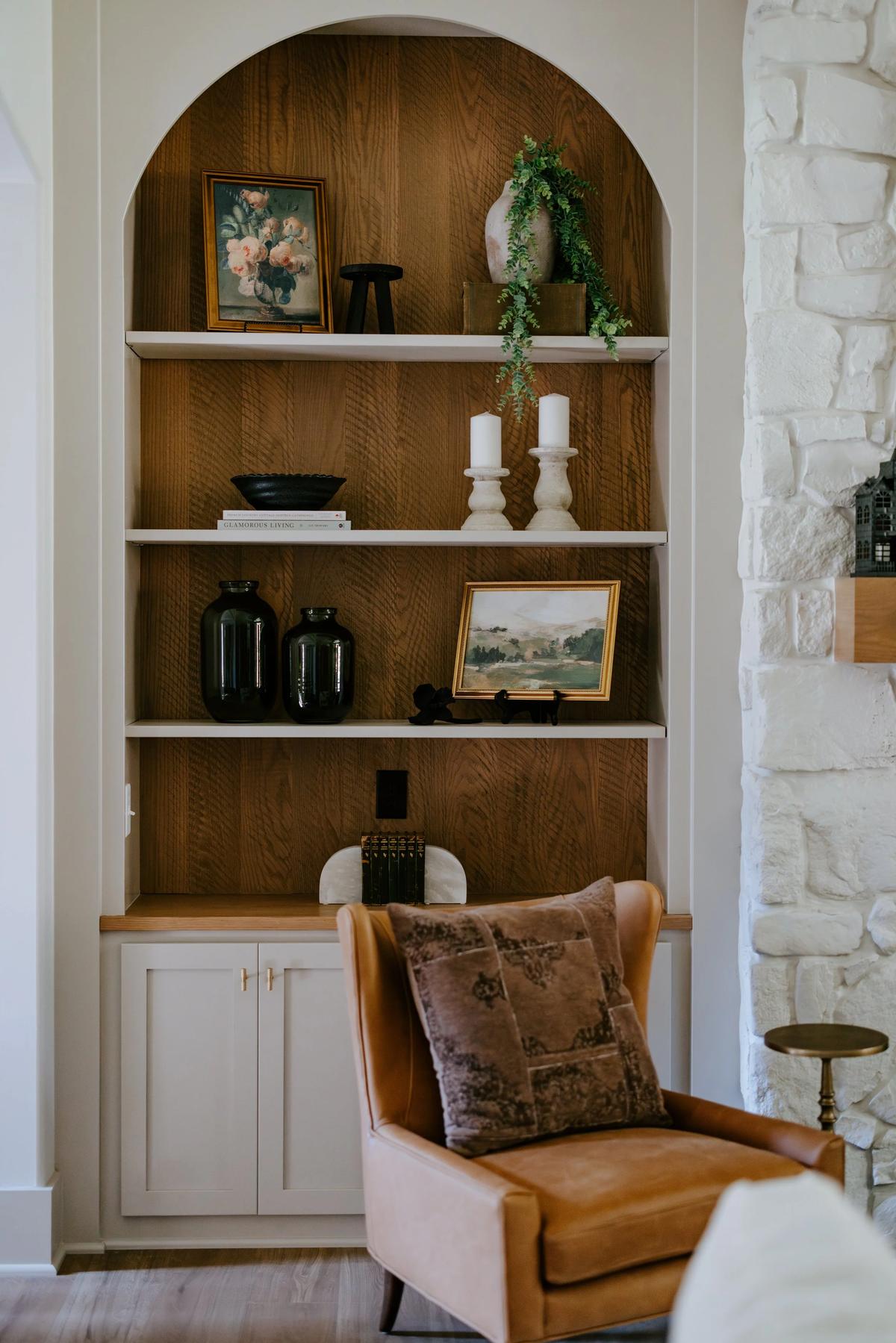 A built-in bookshelf with decorative items, framed artwork, candles, and vases, next to a tan armchair with a velvet pillow in a cozy living room.