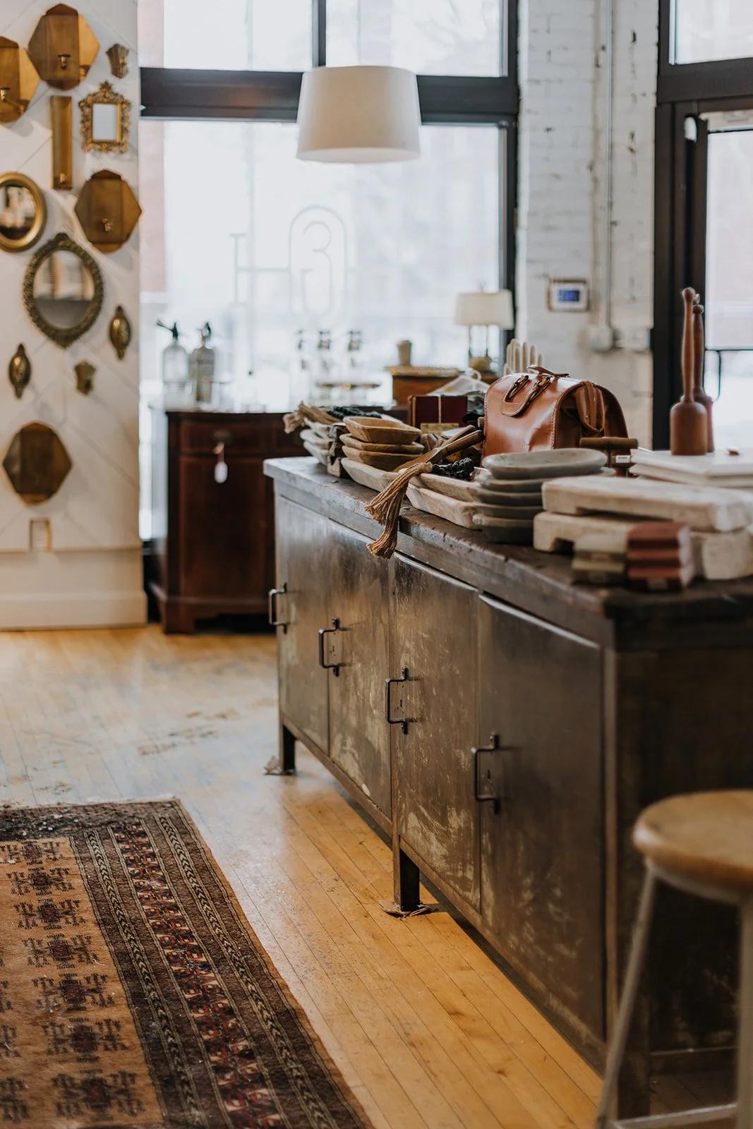 A rustic store interior with a large metal cabinet or counter topped with various decorative plates, bags, and other items. There are vintage mirrors and frames on the white wall to the left, a window letting in natural light, and hardwood flooring with a patterned rug.