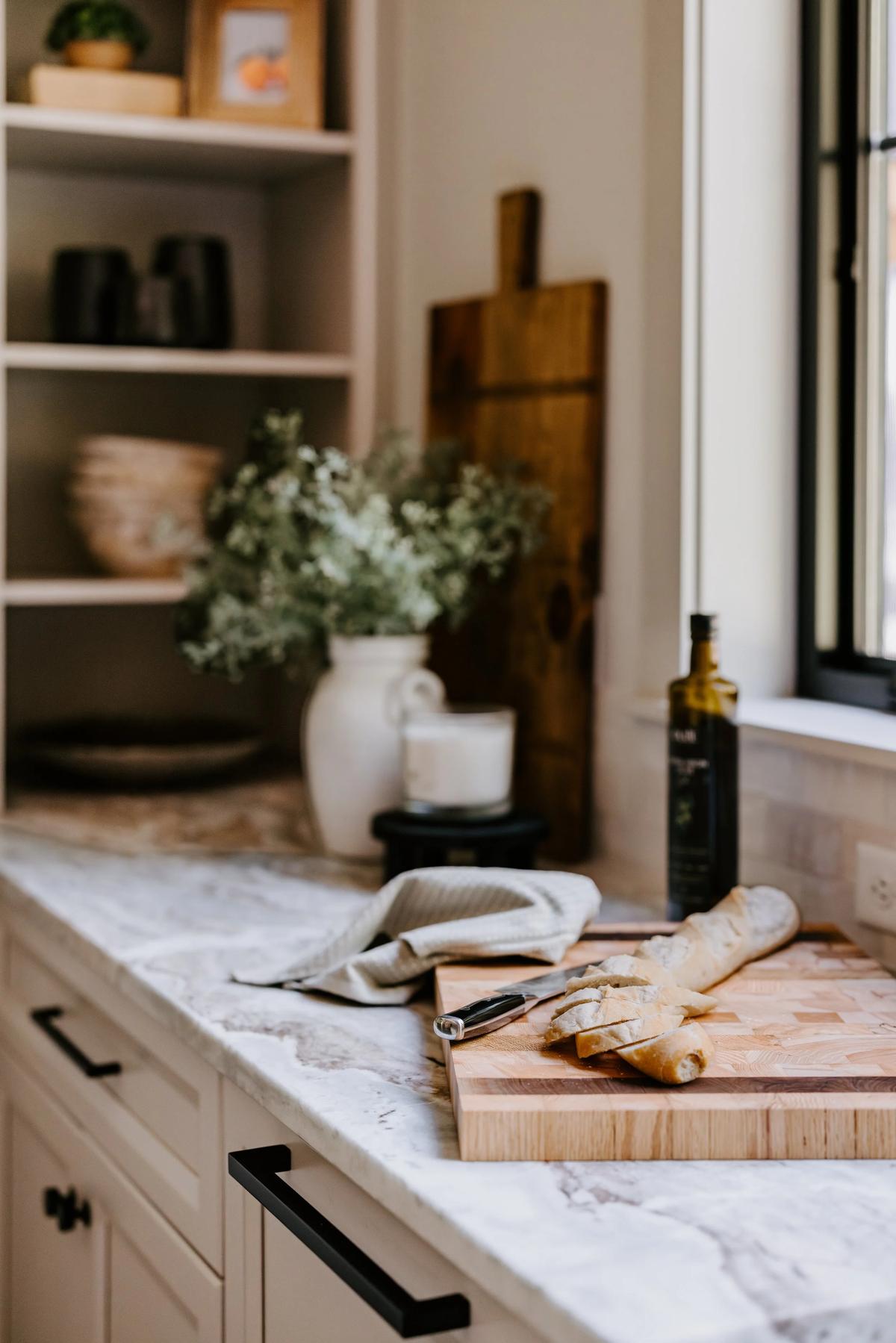 Kitchen countertop with a sliced baguette, a knife, a dish towel, a bottle of olive oil, and decorative items including a white vase with flowers, a candle, and a wooden cutting board.