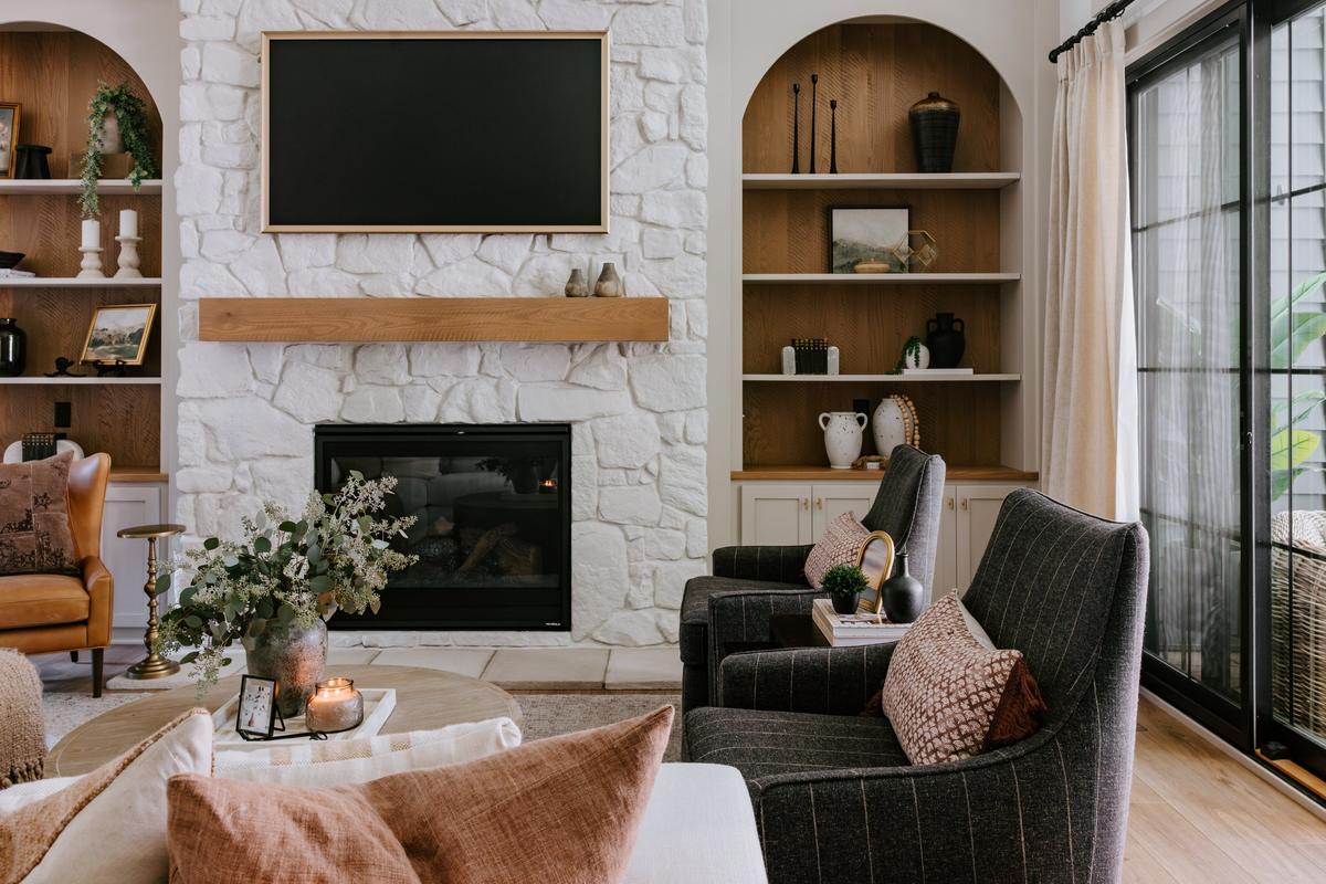 Living room with a white stone fireplace, built-in wooden shelves, and a large window with curtains. There are armchairs, a coffee table with a vase of flowers and candles, and decorative items on the shelves.
