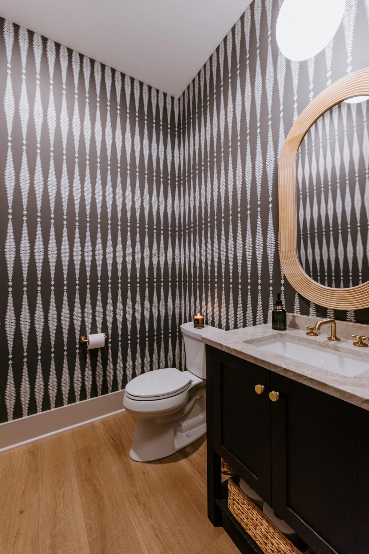 Modern bathroom with black and white patterned wallpaper, a white toilet, and a black vanity with a marble countertop and a large oval mirror.