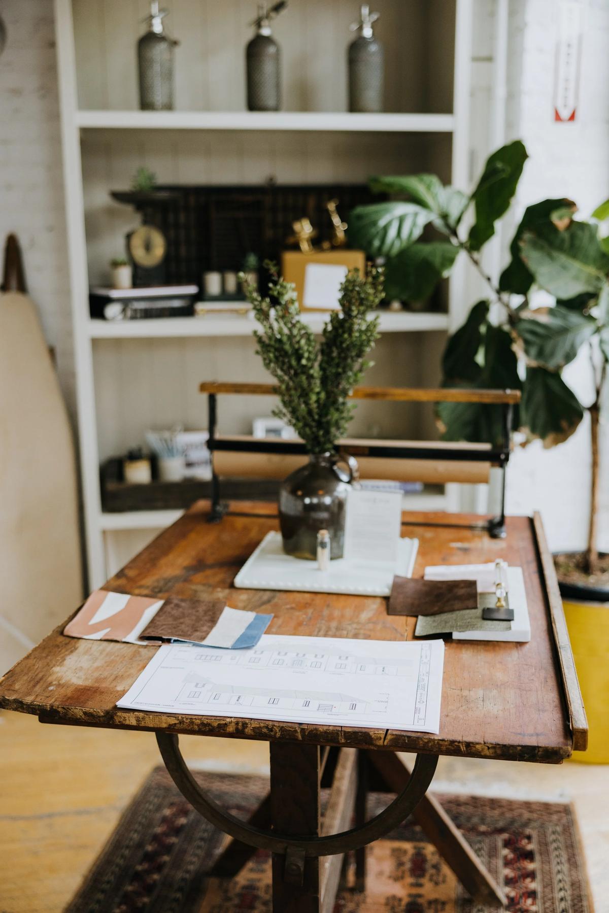 A rustic wooden table with a potted plant and fabric swatches, in a cozy, well-decorated room with a bookshelf and large indoor plant in the background.