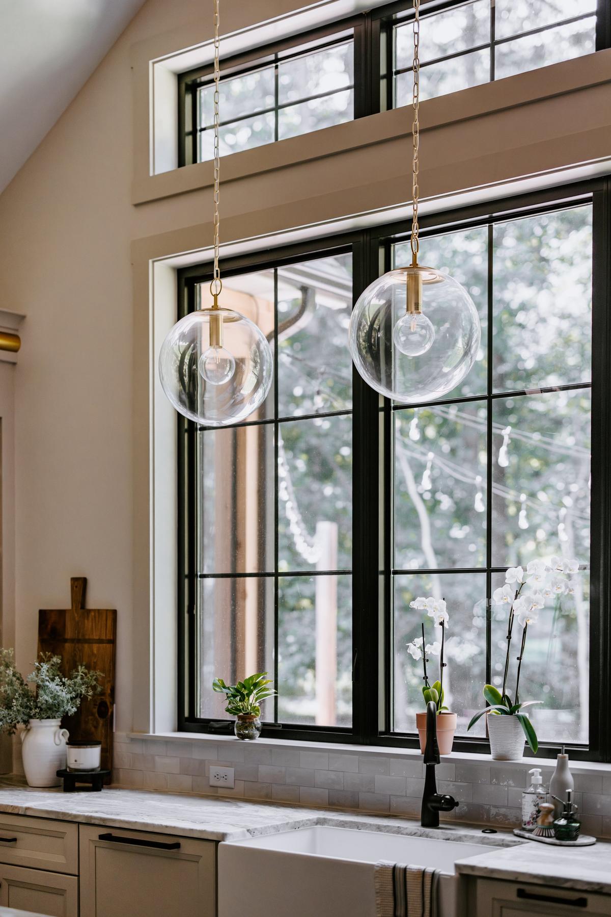 Two glass globe pendant lights hanging over a kitchen sink with a view of a window with black framing outside.