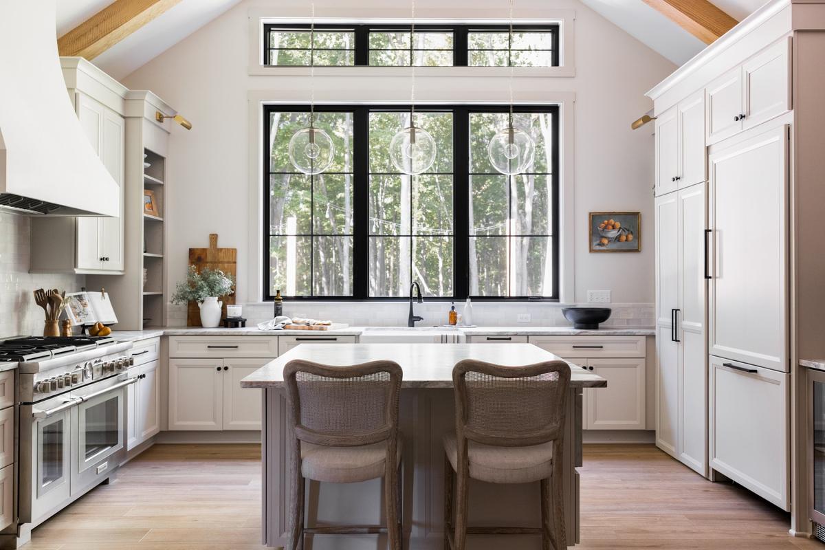 Modern kitchen with white cabinets, island, large black-framed window, pendant lights, and wood accents, with a view of trees outside.
