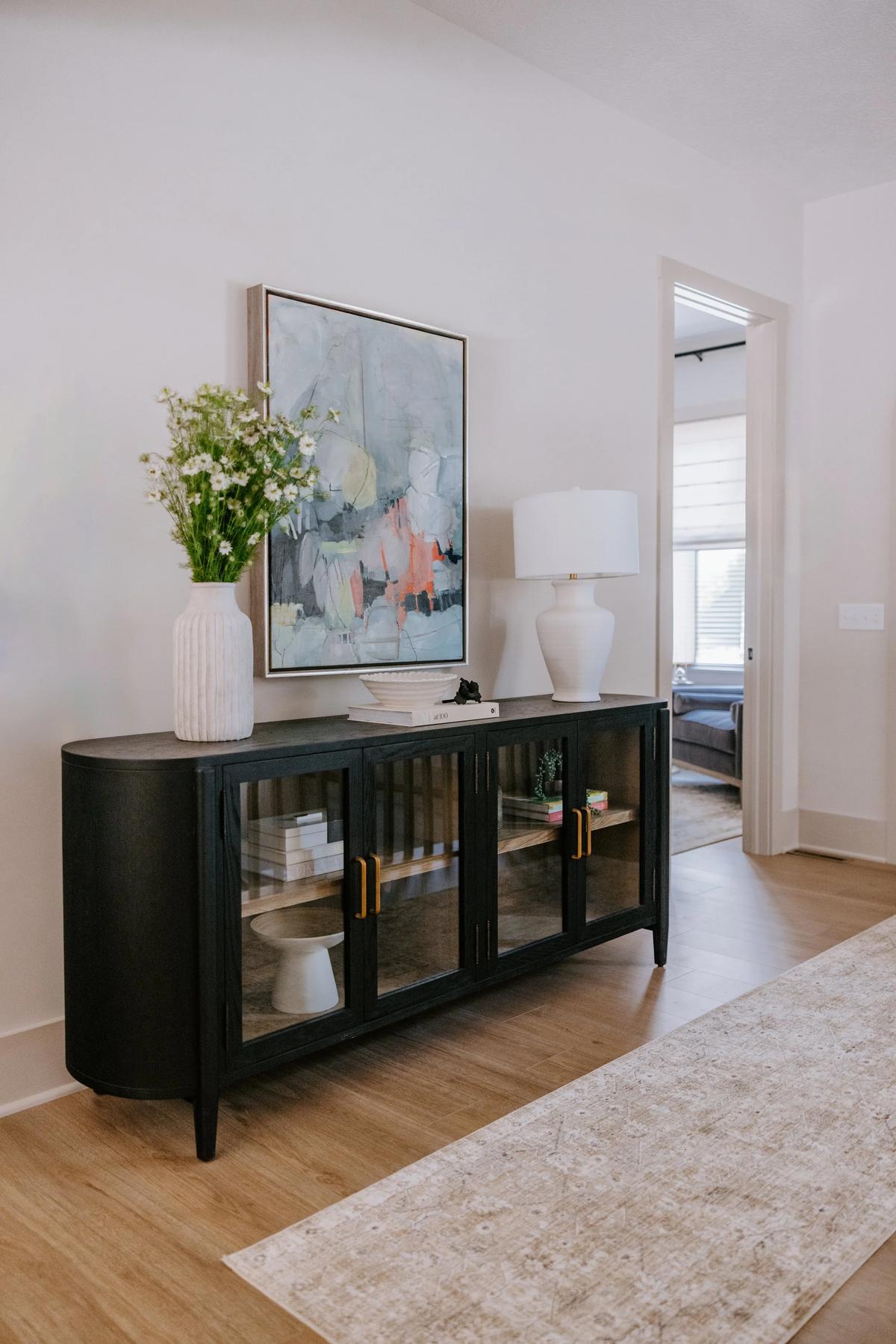 A black glass-front cabinet in a home interior with a white vase of daisies, a white ceramic table lamp, and a stack of books on top, with a framed abstract painting on the wall behind.