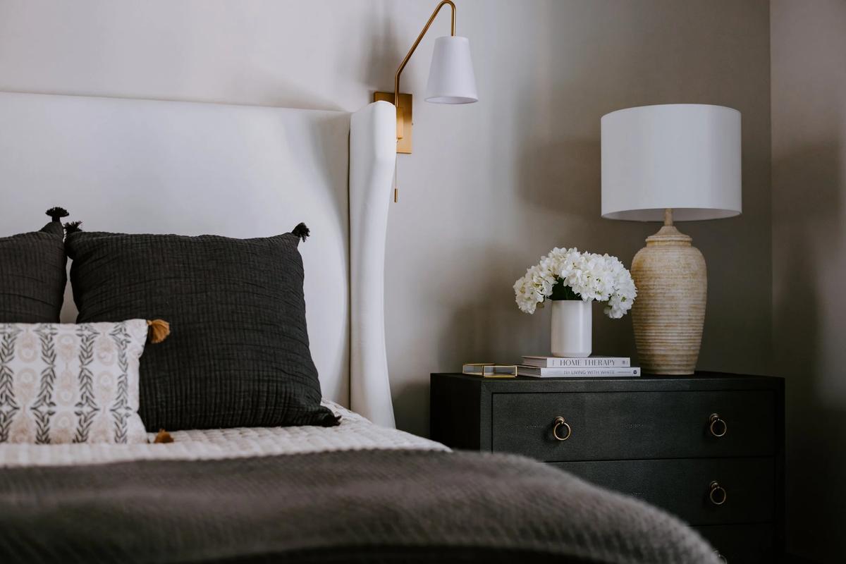 A bedroom with a white headboard, black pillows with tassels, a nightstand with a white flower vase, a large ceramic table lamp, and books.