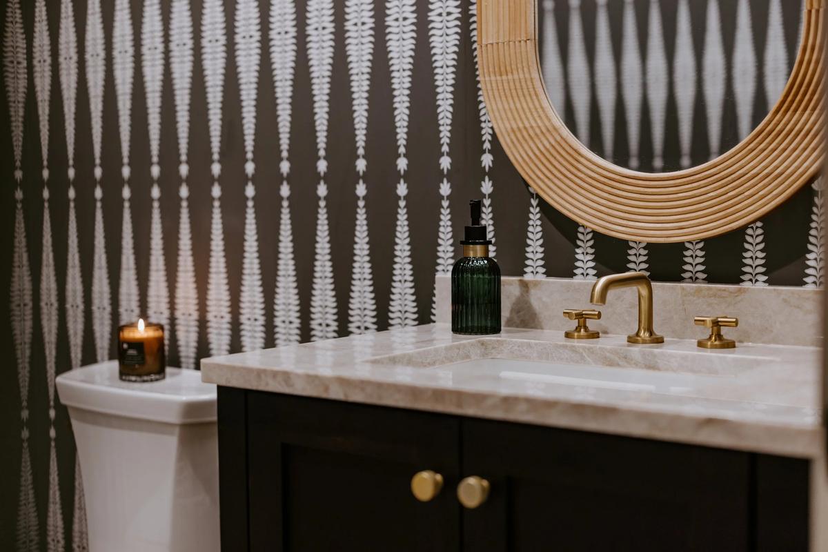 A bathroom vanity with a marble countertop, gold fixtures, a green soap dispenser, and a round mirror, with a lit candle on the toilet tank and patterned wallpaper in the background.