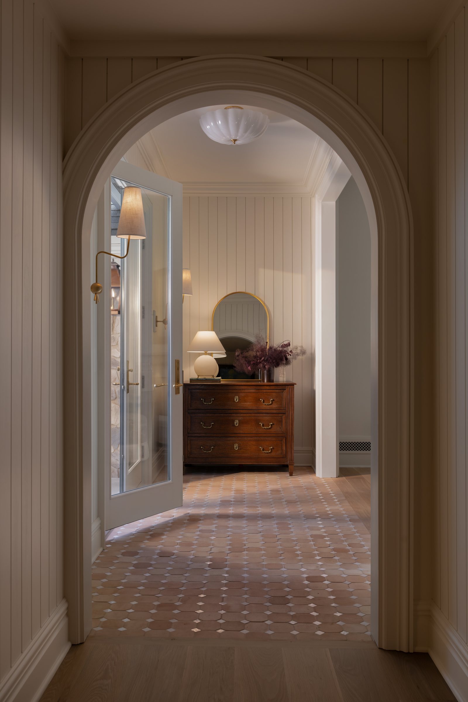 A plaster arch opening onto a panelled entry hall, terracotta and stone floor tile underfoot and a walnut chest beyond