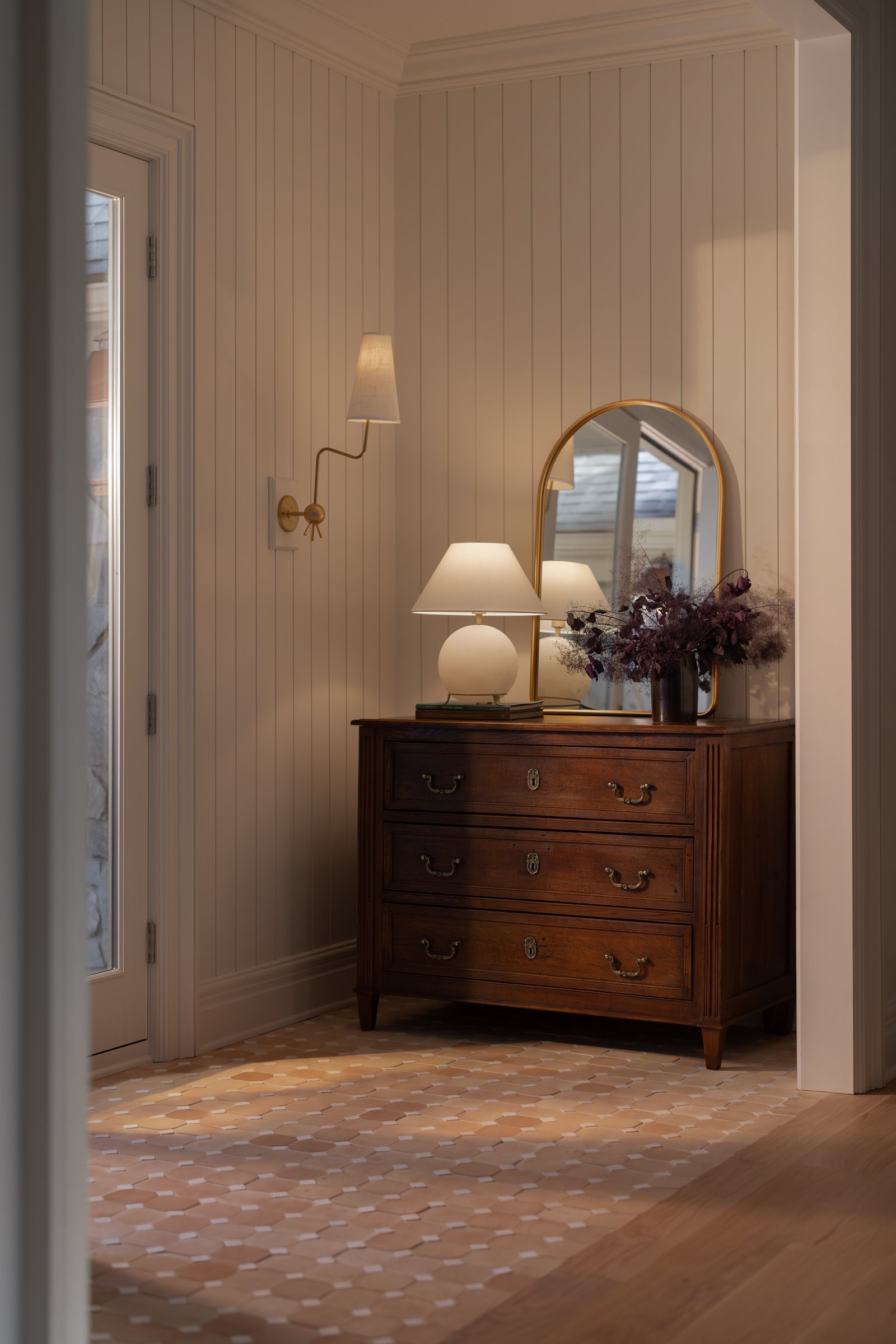 A walnut chest beneath an arched brass mirror, with a globe lamp, books and dried branches, against tongue-and-groove panelling