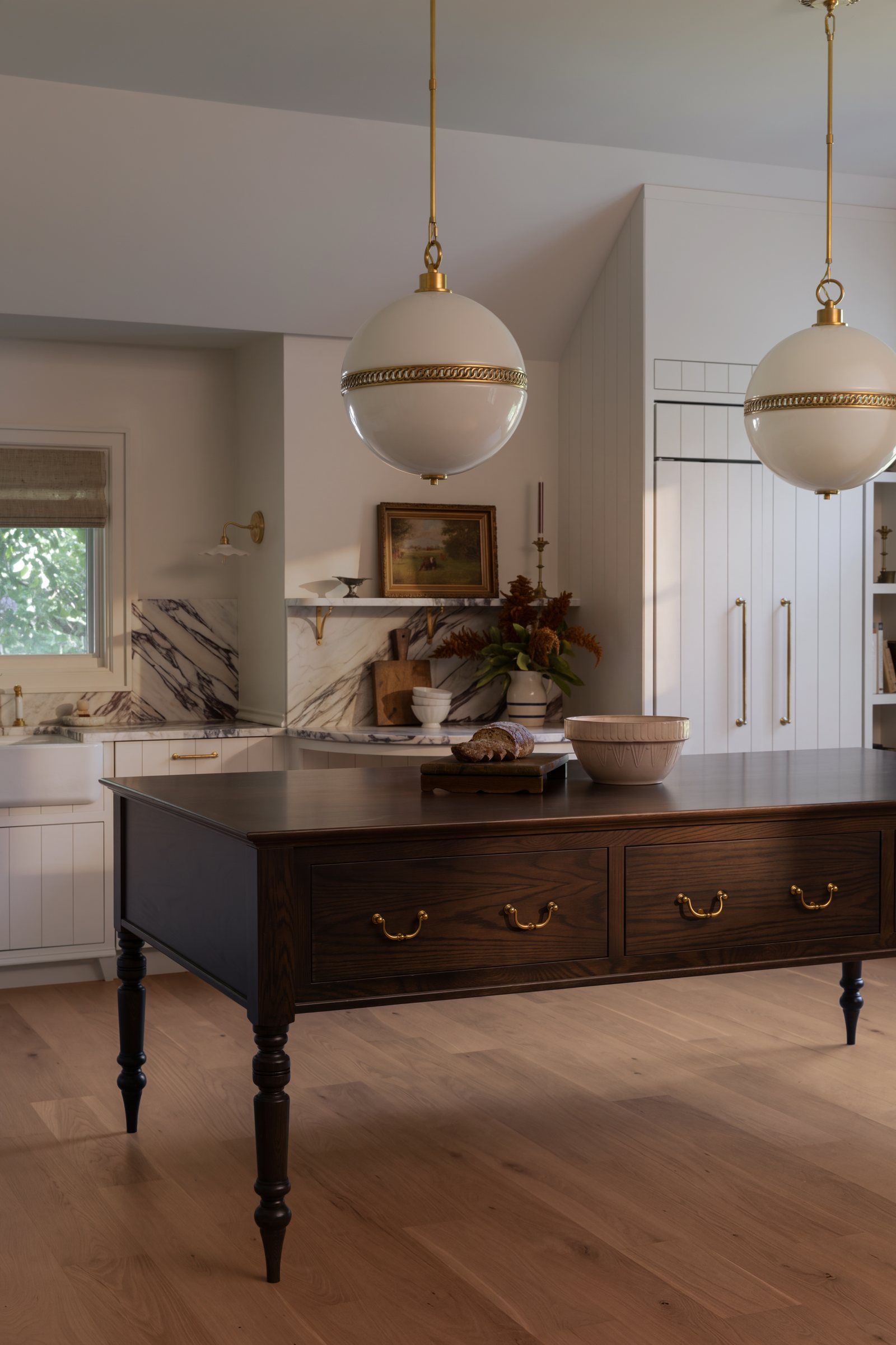 A kitchen with a dark oak island on turned legs, twin globe pendants and a veined marble backsplash