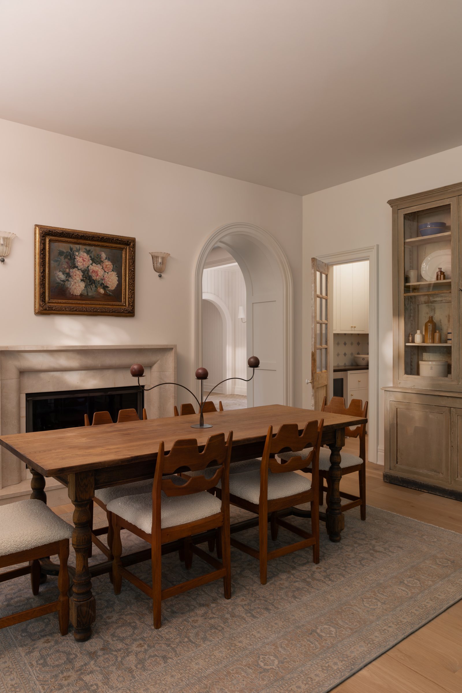 A dining room with an oak trestle table, sculpted chairs and a limestone fireplace, seen beside a plaster arch