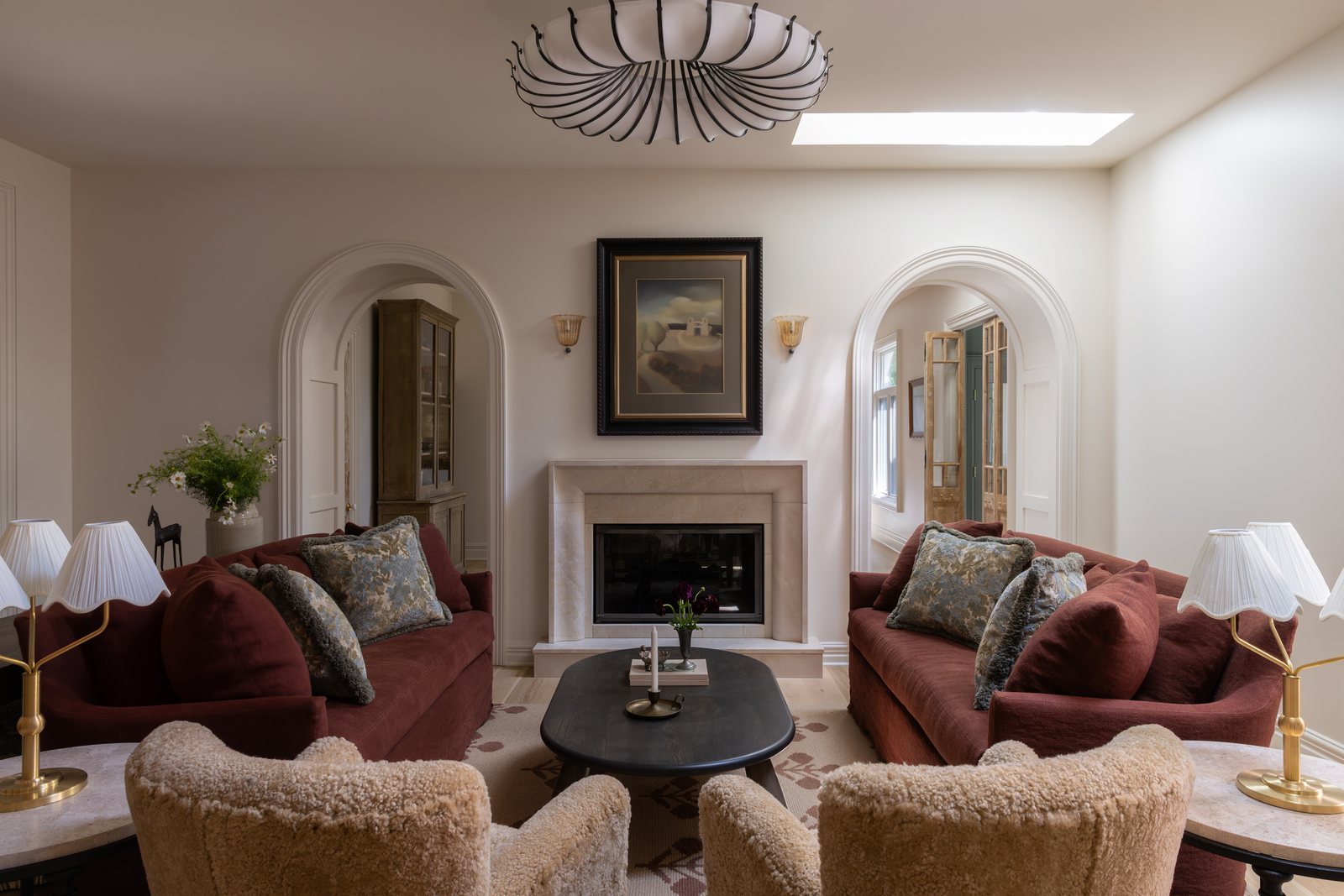 A living room with two deep red sofas facing a limestone fireplace, shearling armchairs and matching plaster arches either side